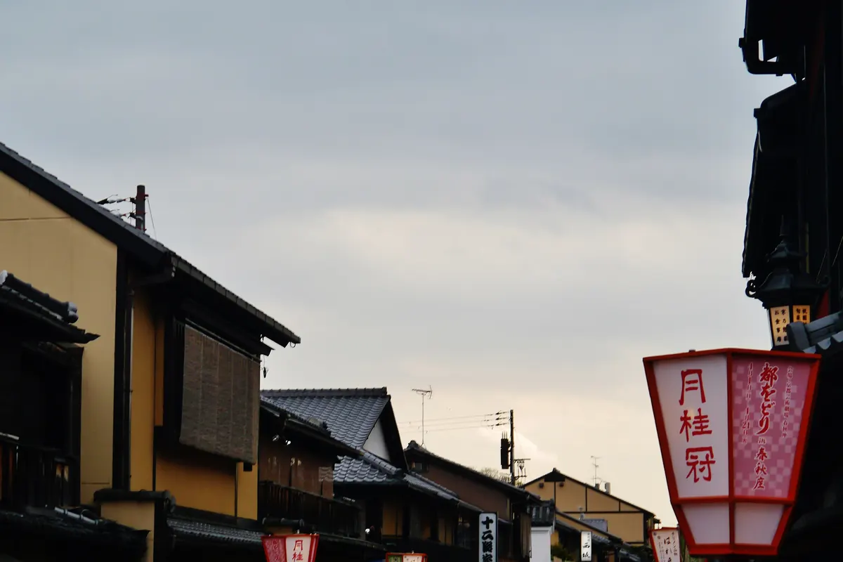 Rote Papierlaternen und dunkle hölzerne Machiya-Häuser entlang einer Straße in Gion, Kyoto, unter abendlichem Himmel
