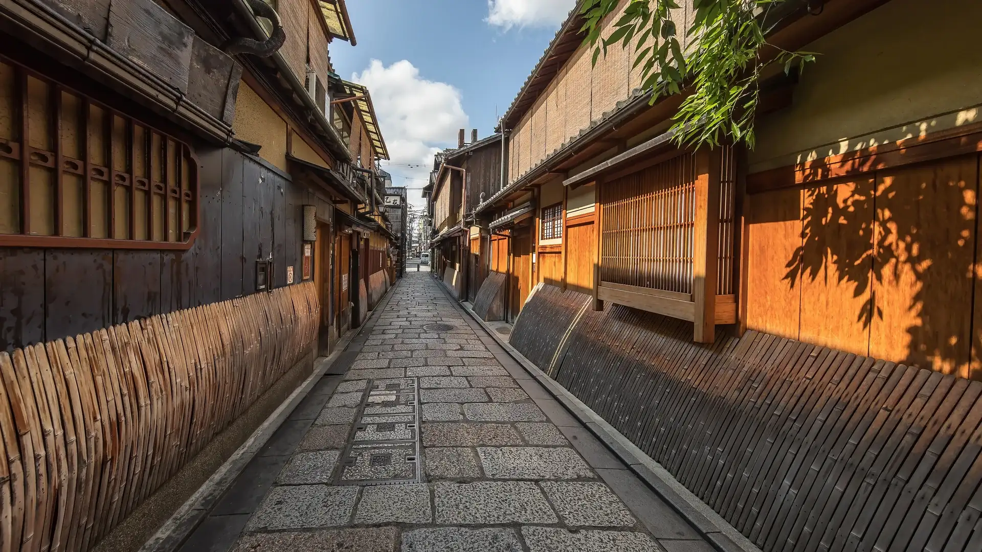 Eine steingepflasterte Gasse in Gion, Kyoto, zwischen hölzernen Stadthäusern mit Bambuszäunen und Lamellenfenstern, in warmem Sonnenlicht