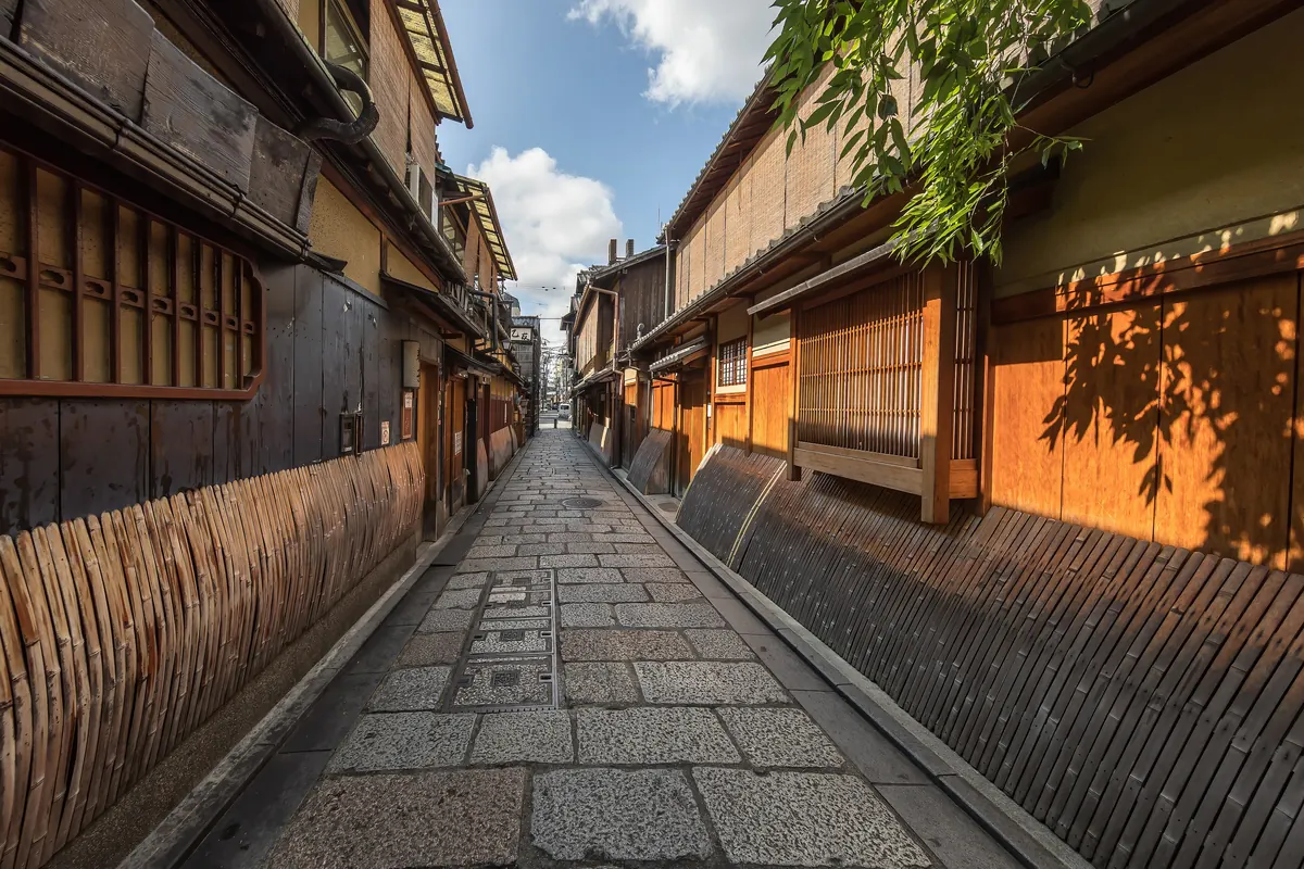 Eine schmale steingepflasterte Gasse in Gion zwischen Holzfassaden mit Bambuszäunen und Lamellenfenstern